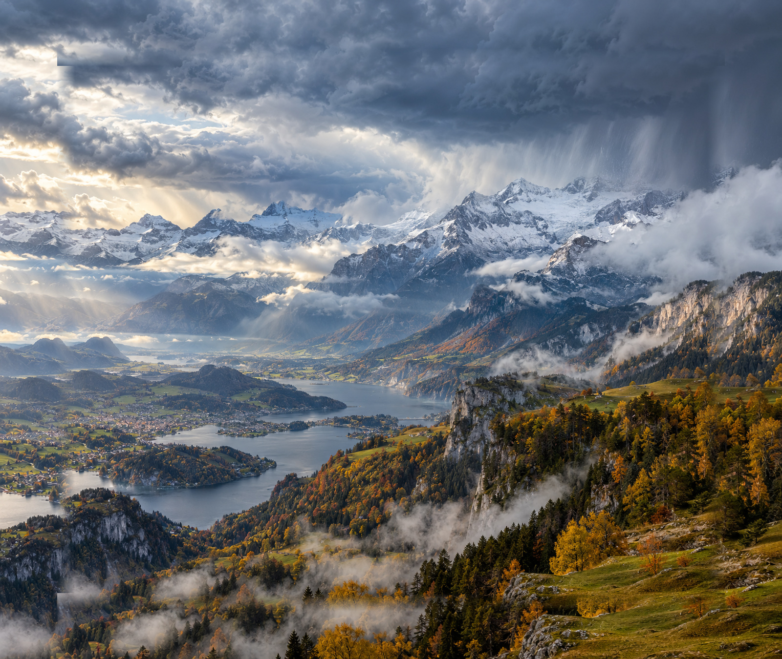 Central European autumn landscape beneath an approaching cold front, with rain clouds and fresh snow visible over the high Alps.